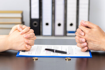 Two people sit across a table in New Jersey, hands clasped. A pen and separation agreement lie between them, with two rings beside the document. Bookshelves with binders blur softly in the background.