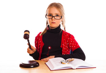 A young girl with glasses, wearing a red polka-dot cardigan, holds a gavel in one hand and is seated at a table. An open book on child custody and a pair of handcuffs lie before her, possibly pondering her future in New Jersey's legal system.
