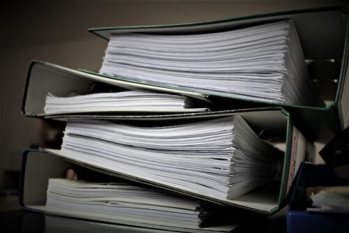 Stack of overflowing binders filled with numerous documents, placed on a dark surface. The binders are slightly open, revealing the clutter of papers inside.